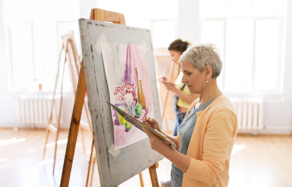 art school, creativity and people concept - senior woman artist with easel, paint brush and palette painting at studio